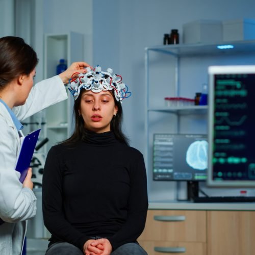Neurological doctor cheking eeg headset explaining to patient diagnosis of disease and treatment for nervous system. Scientist researcher analysing brain scan and tomography on monitor in lab